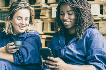 Happy African American and Caucasian factory colleagues in overalls watching content on cellphone...