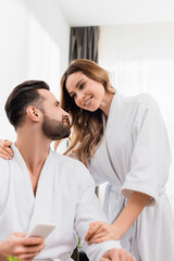 Obraz na płótnie Canvas Smiling woman in bathrobe hugging bearded boyfriend with smartphone on blurred foreground in hotel