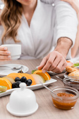Obraz na płótnie Canvas Cropped view of man taking orange slice near food and girlfriend on blurred background in hotel