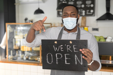 An multiracial waiter, a cafe or bakery owner in protective medical mask is pointing finger at open sign board in his hand. Small business reopening after lockdown