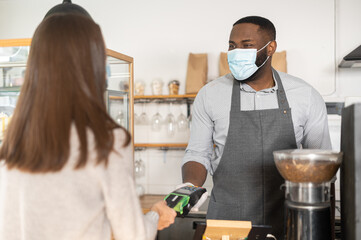 Back view a female customer making contactless payment with a credit card, an African waiter stands behind counter in mask and gloves and holds a terminal. Protective measures during quarantine covid
