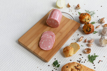 Doctor sausage on wooden cutting board with vegetables, home made bread, spices and greens on white textured background with copy space.