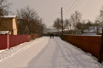 Two people walk along a snow-covered rural street.