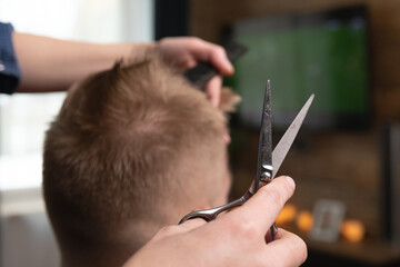 Wife cutting husband's hair at home in front of TV . Scissors in focus.
