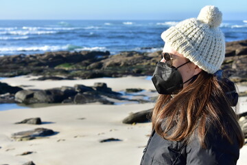 Fototapeta na wymiar Woman with Covid-19 black face mask on a beach in Winter with black coat, sunglasses and white wool hat. KN95 or N95 or FFP2 mask.