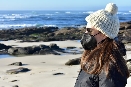Woman with Covid-19 black face mask on a beach in Winter with black coat, sunglasses and white wool hat. KN95 or N95 or FFP2 mask.