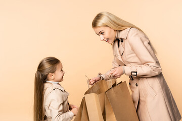 smiling mother holding paper bags near cheerful daughter isolated on beige
