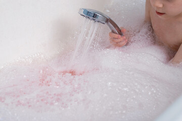 Fototapeta premium The child makes lather in the bath with the shower. Taking a bath, hygiene procedures.