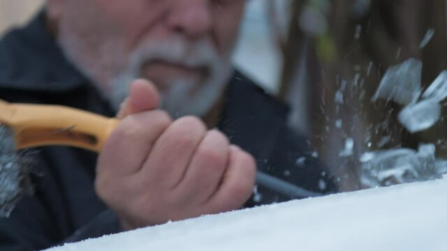 Senior person with grey beard hits automobile roof with black rubber ice scraper on cold winter day