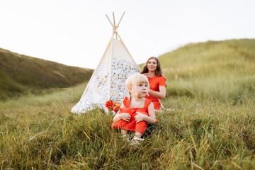 Beautiful mother with her little daughter sitting near wigwam in the field. Spending time together, outside, on vacation, outdoors. Beautiful sunset light in the garden or in the park © Andriy Medvediuk