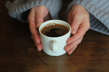 Woman's hands in sweater hold a cup of strong coffee on wooden table. Coffee fan top view background