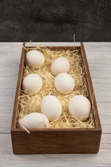 Seven white chicken eggs are lying in a wooden box on the straw
