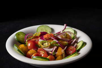 Fresh two-color cherry tomato salad, cucumber, red onion and hot chili served in a vintage bowl on a table. 45 deg. view.Black background.