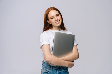Charming young woman student holding laptop computer and looking at camera on isolated gray background. Pretty lady model with red hair emotionally showing facial expressions in studio, copy space.