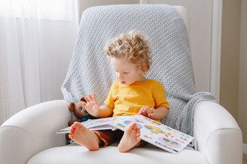 Cute adorable little baby boy toddler 2 years old sitting in armchair at kids room and reading book. Early age kid child development, education. Candid home authentic childhood lifestyle. - 407052517
