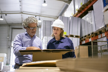 Focused safety inspector consulting female engineer while completing documents during inspection of warehouse. Front view. Labor and corporate communication concept