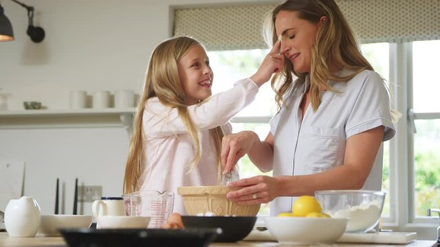 Mother and daughter wearing pyjamas in kitchen putting mixture on each other's noses as they bake and make pancakes at counter together - shot in slow motion - Powered by Adobe