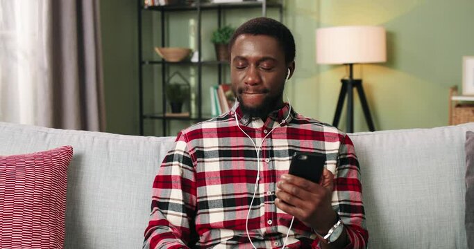 Portrait of handsome cheerful young African American guy enjoying music in earphones on cellphone listening to fav songs resting on couch in cozy apartment alone. Home concept, leisure time