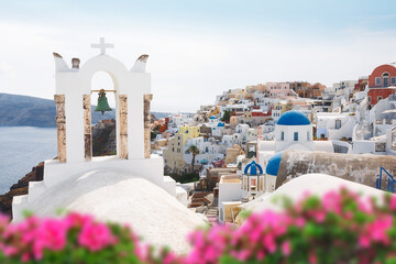 Oia village  through flowers and  traditional Greek white church arch with cross and bells in Oia village of Cyclades Island, Santorini