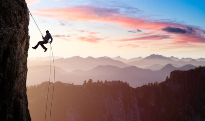 Adventurous Man Rappelling from Cliff. Beautiful aerial view of the mountains during a colorful and...