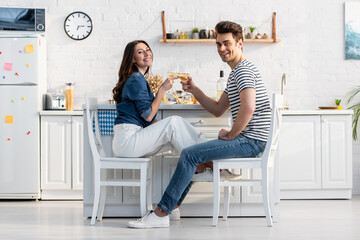 happy couple holding glasses with wine and clinking while sitting in kitchen