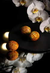 Easter eggs lie on a slate on a mirrored table next to orchid flowers.  Easter day. Easter eggs with marble coloring. Marble coloring. Dark background.