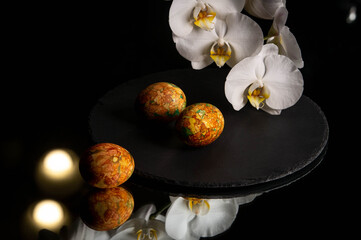 Easter eggs lie on a slate on a mirrored table next to orchid flowers.  Easter day. Easter eggs with marble coloring. Marble coloring. Dark background.