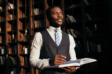 Stylish attractive male African American teacher at the university stands with a magazine on the background of shelves with books.