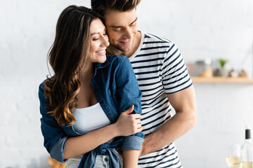 Naklejka premium happy man embracing joyful girlfriend and smiling in kitchen