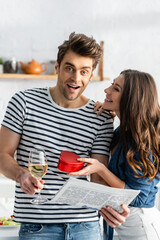 amazed man holding glass of wine and newspaper near cheerful girlfriend with heart-shaped gift box