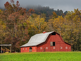Fog lays over a green field and red country barn in the fall season.