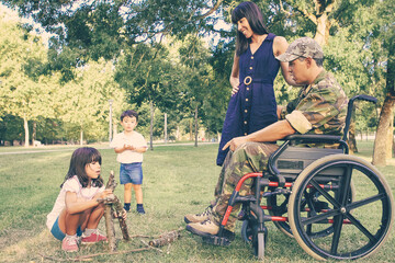 Fototapeta premium Cheerful mom and disabled military dad in wheelchair looking at kids arranging firewood for campfire outdoors. Disabled veteran or family outdoors concept