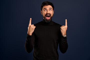 Horizontal portrait of bearded cheerful man has smile, wears trendy sweater, indicates with fingers at copy space for your promotional text or advertisment, isolated over Pacific Blue background