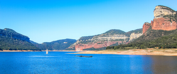 Panoramic mountainous landscape of the Sau swamp with its church. Tavertet and Collsacabra cliffs in the Sau Reservoir on a sunny day. Tourism in Osona, Barcelona, Catalonia, Spain.