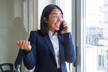 Cheerful Latin office employee talking on cellphone, speaking and smiling, standing at window in office . Communication concept