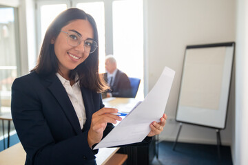 Happy female professional holding documents and pen, looking at camera and smiling. Medium shot. Business and paperwork concept