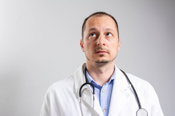 Aged friendly doctor looks up to adspace and smiles.Isolated on grey background. Portrait of male doctor with stethoscope in medical uniform looking up posing on a grey isolated background.