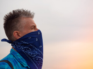 Portrait of a caucasian man in the rays of the setting sun. His face covered with a blue bandana,...