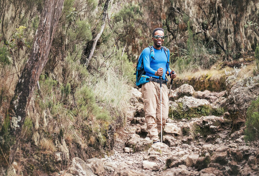 Portrait of a cheerfully smiling African-American Ethnicity young man in sunglasses. He having a walk with a backpack using trekking poles to Mweka gate, Kilimanjaro. Active climbing people concept.
