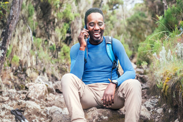 Naklejka na ściany i meble Young African ethnicity man talking via cellphone with somebody and cheerfully laughing as he having a hiking walk in the tropical forest. Happy people, network technology, or traveling concept.
