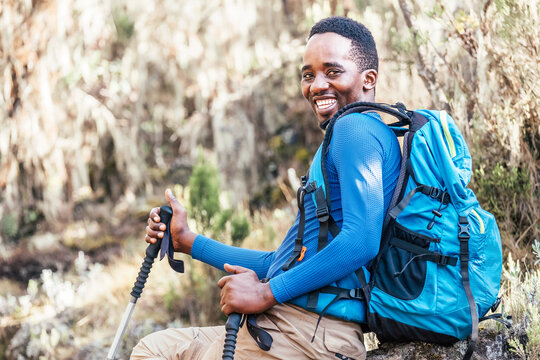 Portrait of a cheerfully smiling African-American Ethnicity young man sitting with a backpack and trekking poles and resting in the forest during a hiking walk. Active people and sport concept.