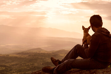 Young man spending time together with his dog pet, retiring in nature in top of hill and watching...