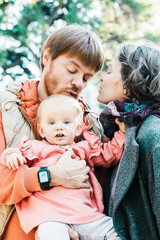 Naklejka premium Cute couple of parents and little daughter posing outdoors. Mom and dad holding cute baby girl in arms, standing in autumn park. Vertical shot. Family portrait concept