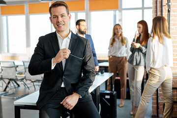 leader of business team in the office, he is posing at camera while colleagues coworking in the background, sits on table