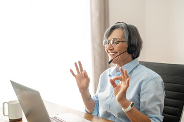 Senior lady call center operator with a grey hair sitting at the desk, wearing glasses and a...