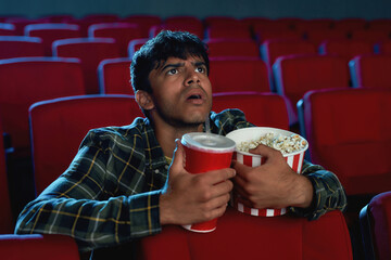 Portrait of concentrated young guy holding a drink and popcorn basket while watching movie alone in empty theater auditorium