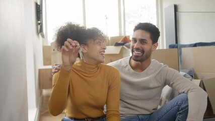 Portrait of young mixed ethnicity couple sitting on floor in lounge holding keys to new home unpacking removal boxes together - shot in slow motion