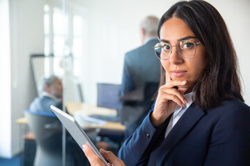 Confident business lady in glasses and suit holding tablet, touching chin and looking at camera. Two businessmen working behind glass wall. Copy space. Communication concept