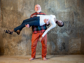 elderly man with gray beard in plaid shirt and red pants lifted beautiful African American girl in his arms and held her. Interracial marriage concept