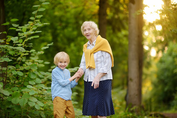 Cute grandson holding hands with his joyful elderly grandmother during walking at summer park.